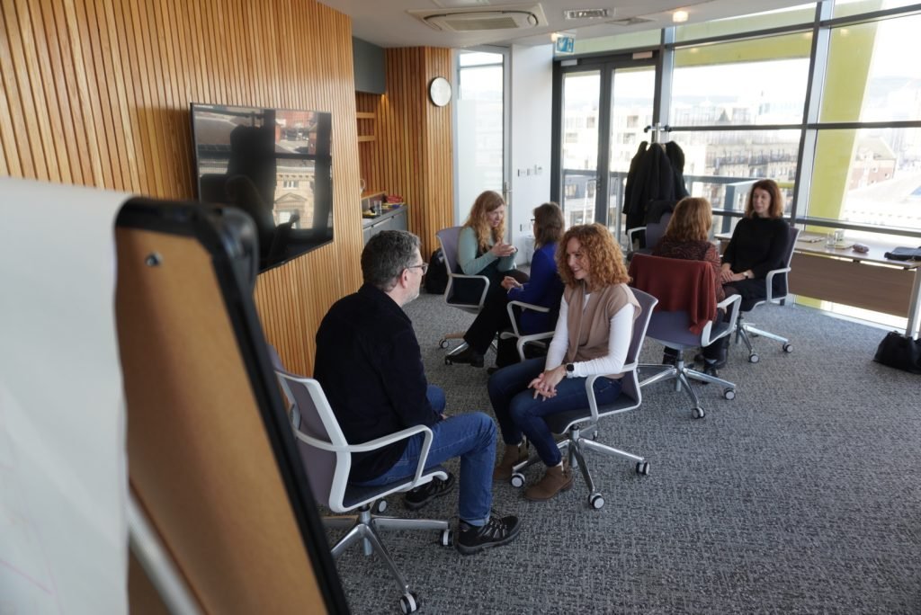 A group of adults seated in chairs, engaged in a calm discussion in a bright, well-lit office space.
