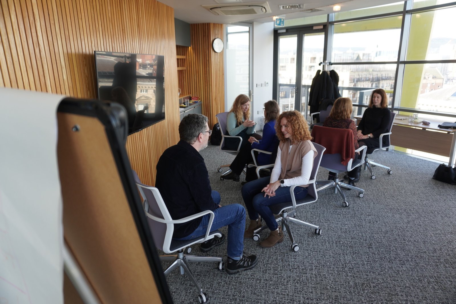 A group of adults seated in chairs, engaged in a calm discussion in a bright, well-lit office space.